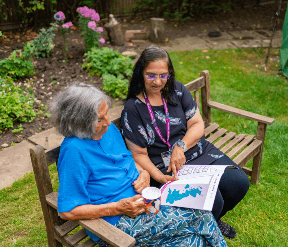 Two women sitting on a wooden bench in a garden, looking at a document with a map. One is holding a mug. - Home Instead