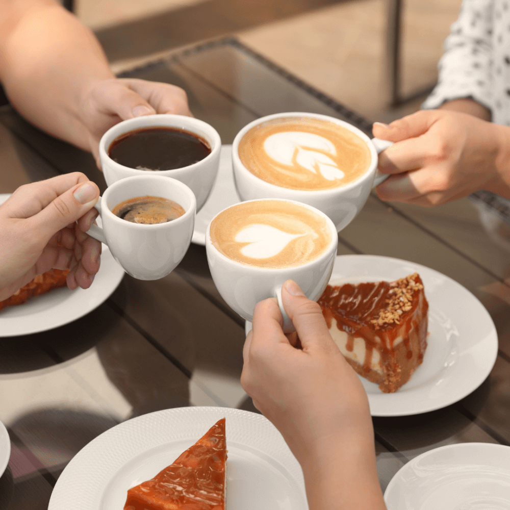 Four hands holding cups of coffee for a toast, surrounded by caramel-topped desserts on a table. - Home Instead