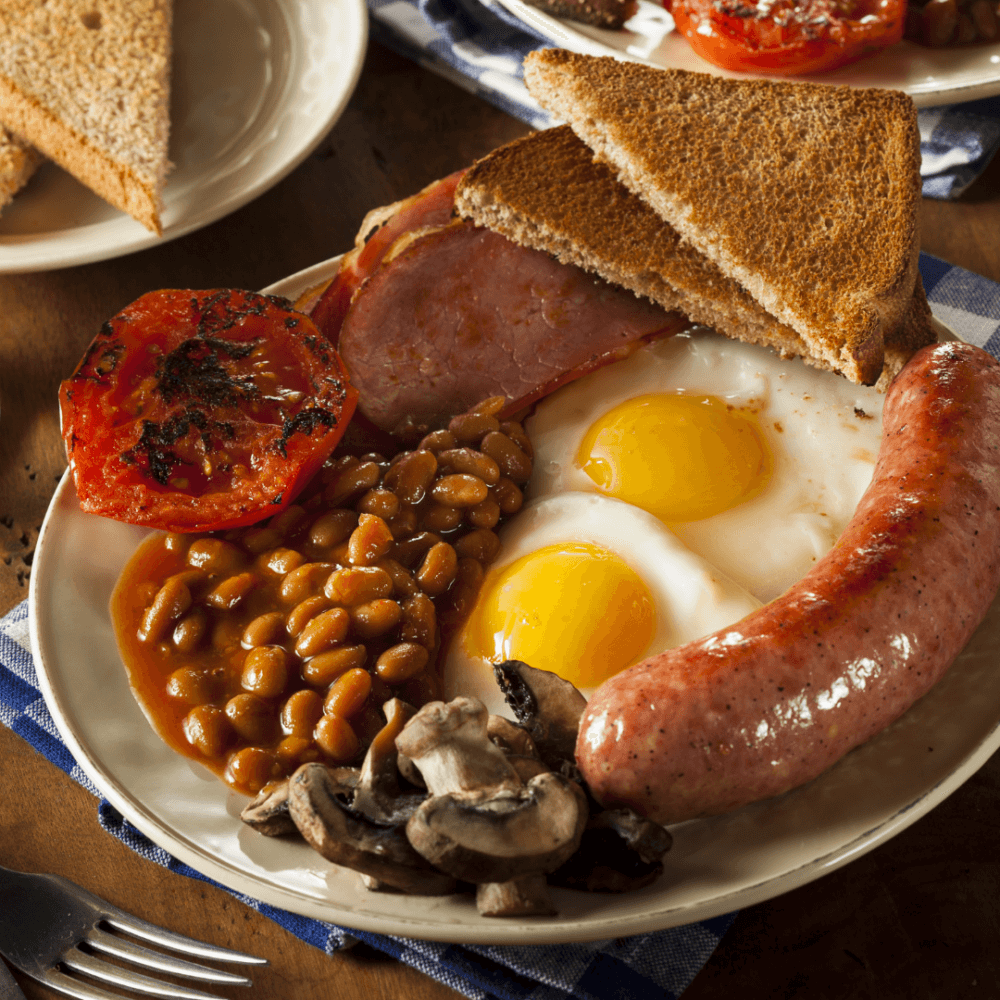 A hearty breakfast of eggs, sausage, bacon, baked beans, grilled tomatoes, mushrooms, and toast on a white plate. - Home Instead