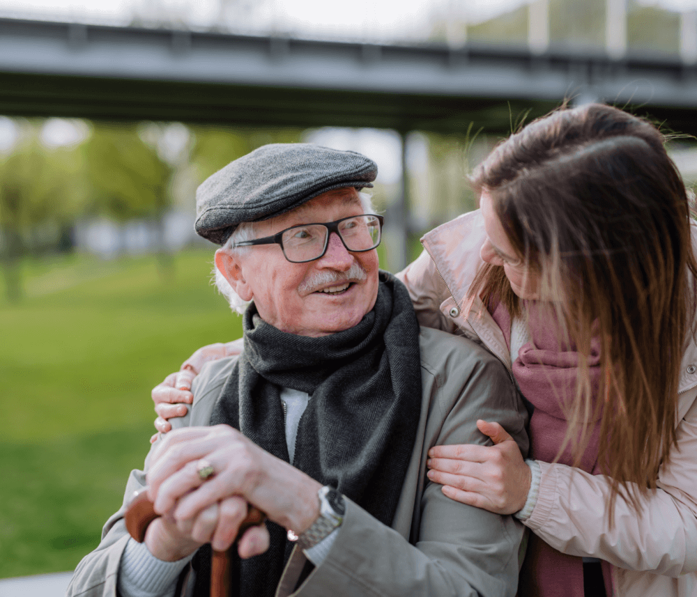 An elderly man with glasses and a flat cap smiles as a young woman in a scarf embraces him in an outdoor park. - Home Instead
