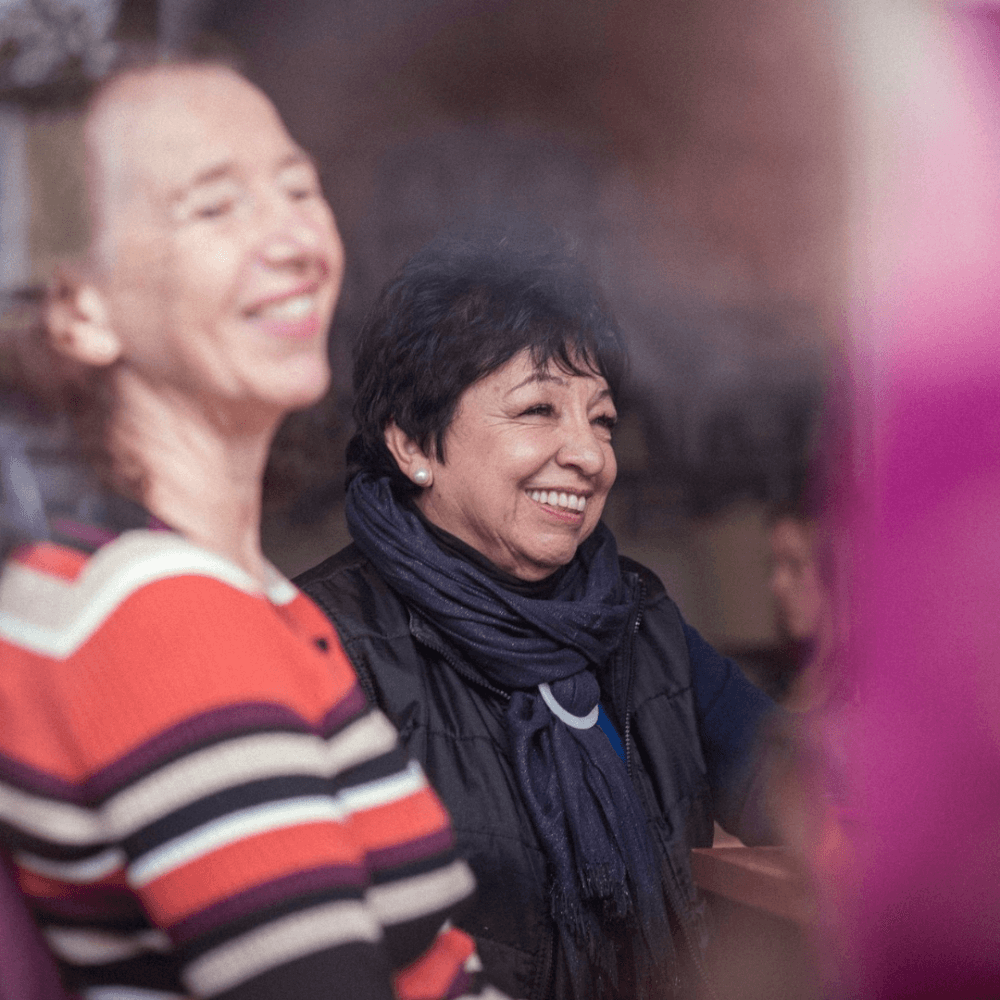 Two women smiling and enjoying a conversation; one in a striped sweater, the other in a dark jacket and scarf. - Home Instead