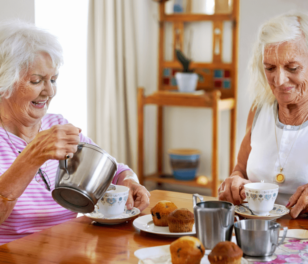 Two elderly women enjoying tea and muffins at a wooden table, one pouring tea while smiling. - Home Instead