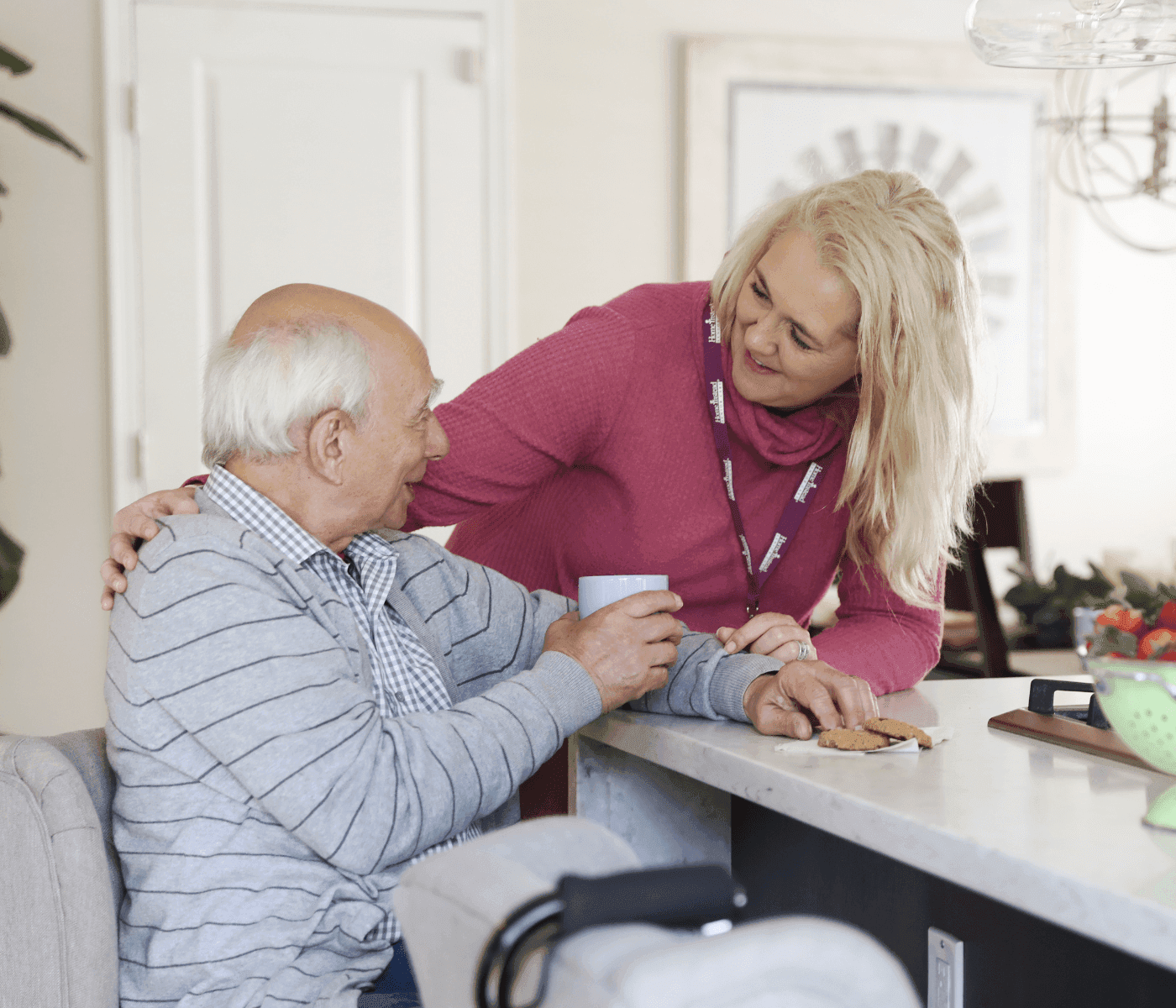 A smiling woman in a pink shirt assists an elderly man seated at a kitchen counter, holding a cup of coffee. - Home Instead