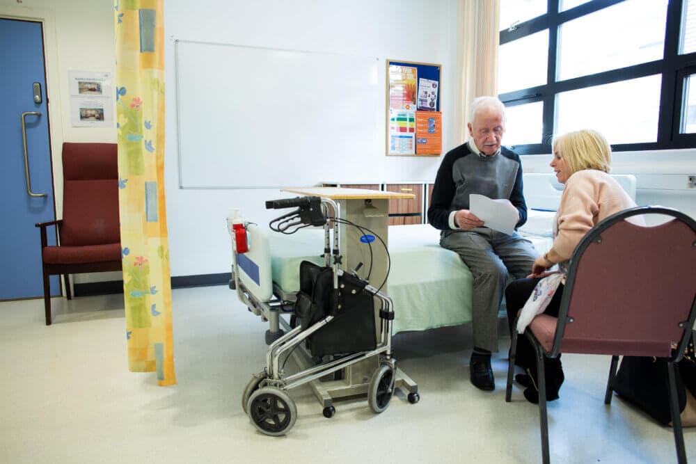 An elderly man and a woman sit beside a hospital bed, holding papers. A wheelchair and medical equipment are nearby. - Home Instead