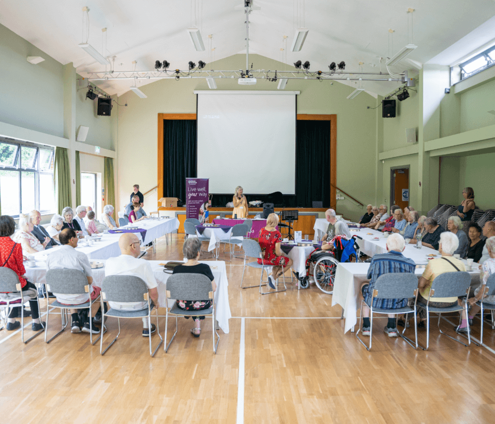 A group of elderly people sit around tables in a bright hall, listening to a speaker at a podium. - Home Instead