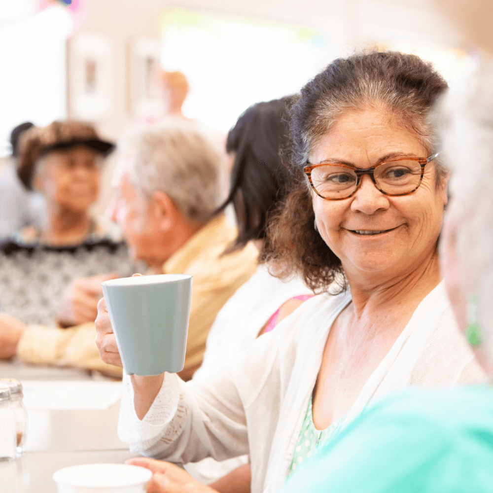 Older adults enjoying a social gathering; a woman in glasses holds a cup and smiles at another person in the foreground. - Home Instead