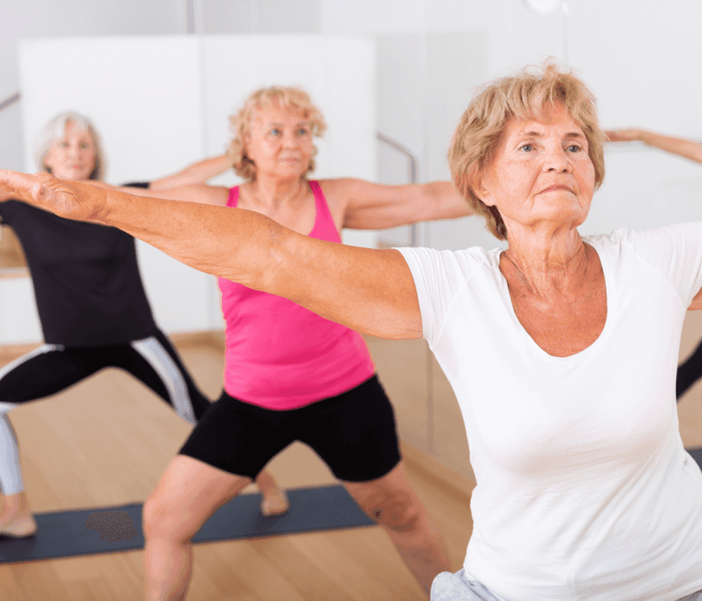 Three senior women practicing yoga in a studio, doing warrior poses on yoga mats. - Home Instead