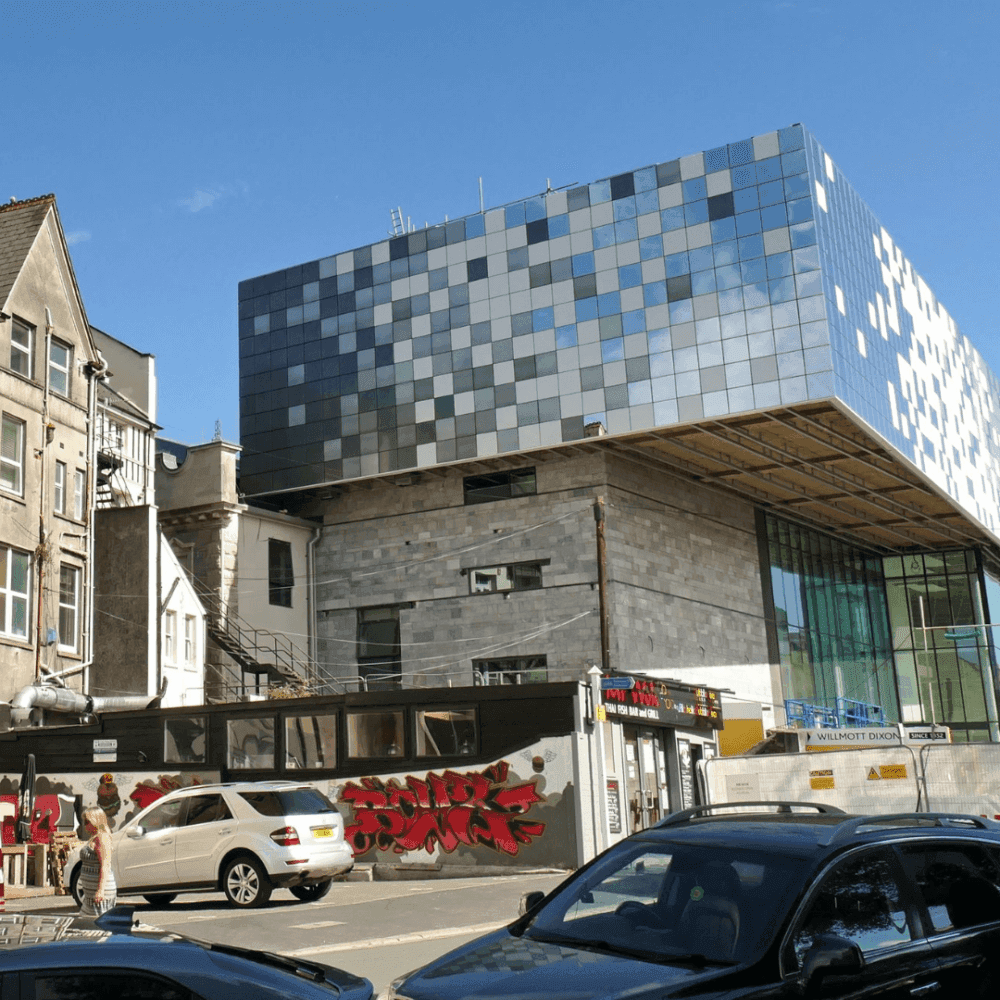 Modern building with a pixelated facade atop traditional structures, surrounded by parked cars and graffiti-covered walls. - Home Instead
