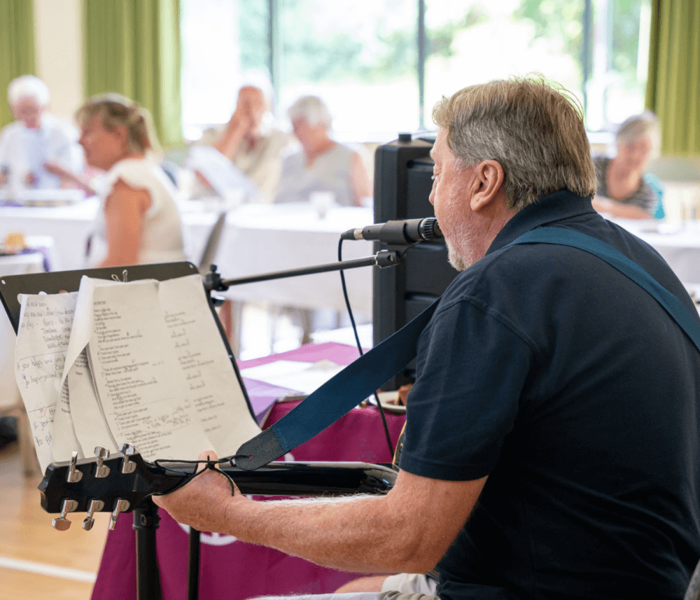 A man with a guitar performs indoors. An audience sits at tables in the background. - Home Instead