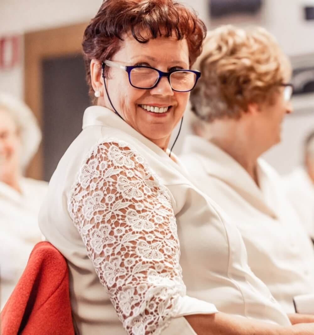 A woman with glasses and a lace-sleeved top smiles while sitting in a group setting. - Home Instead