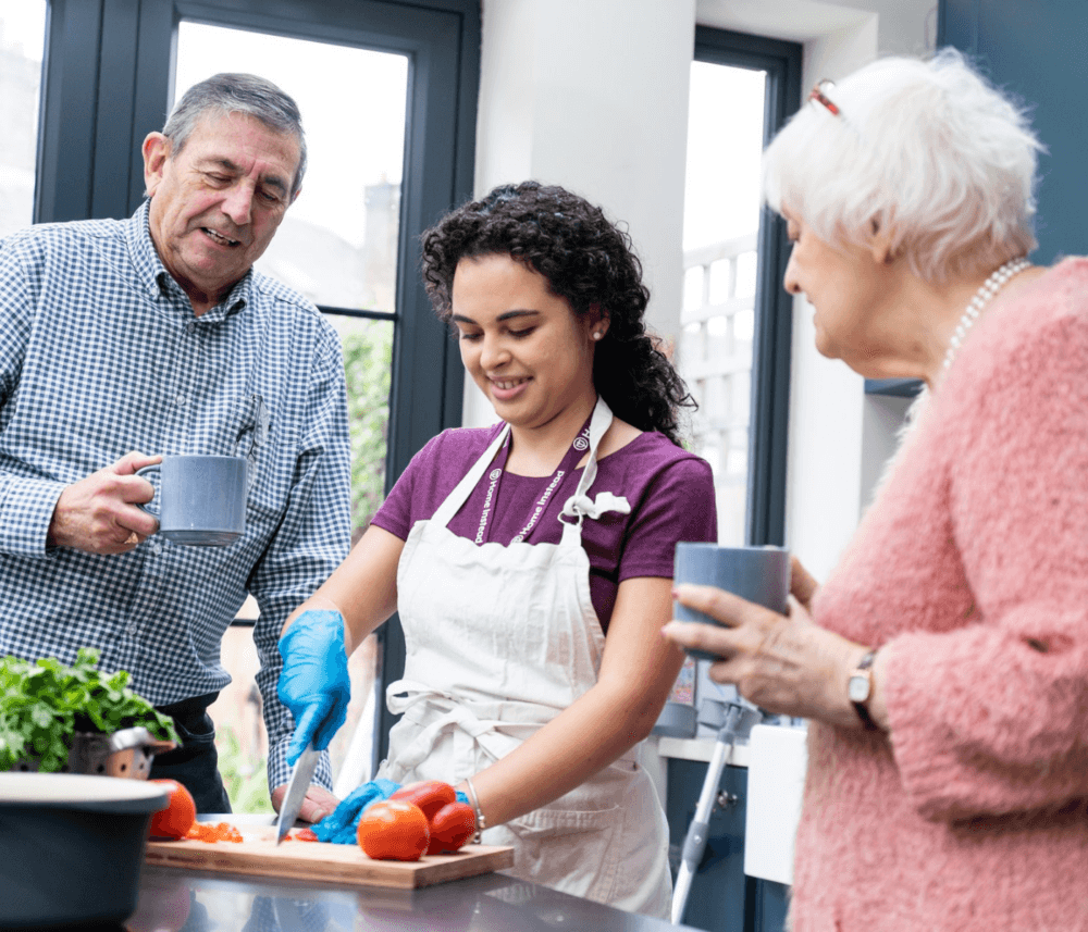 Young woman prepares food at kitchen counter as two older adults watch, all holding mugs and enjoying their time together. - Home Instead