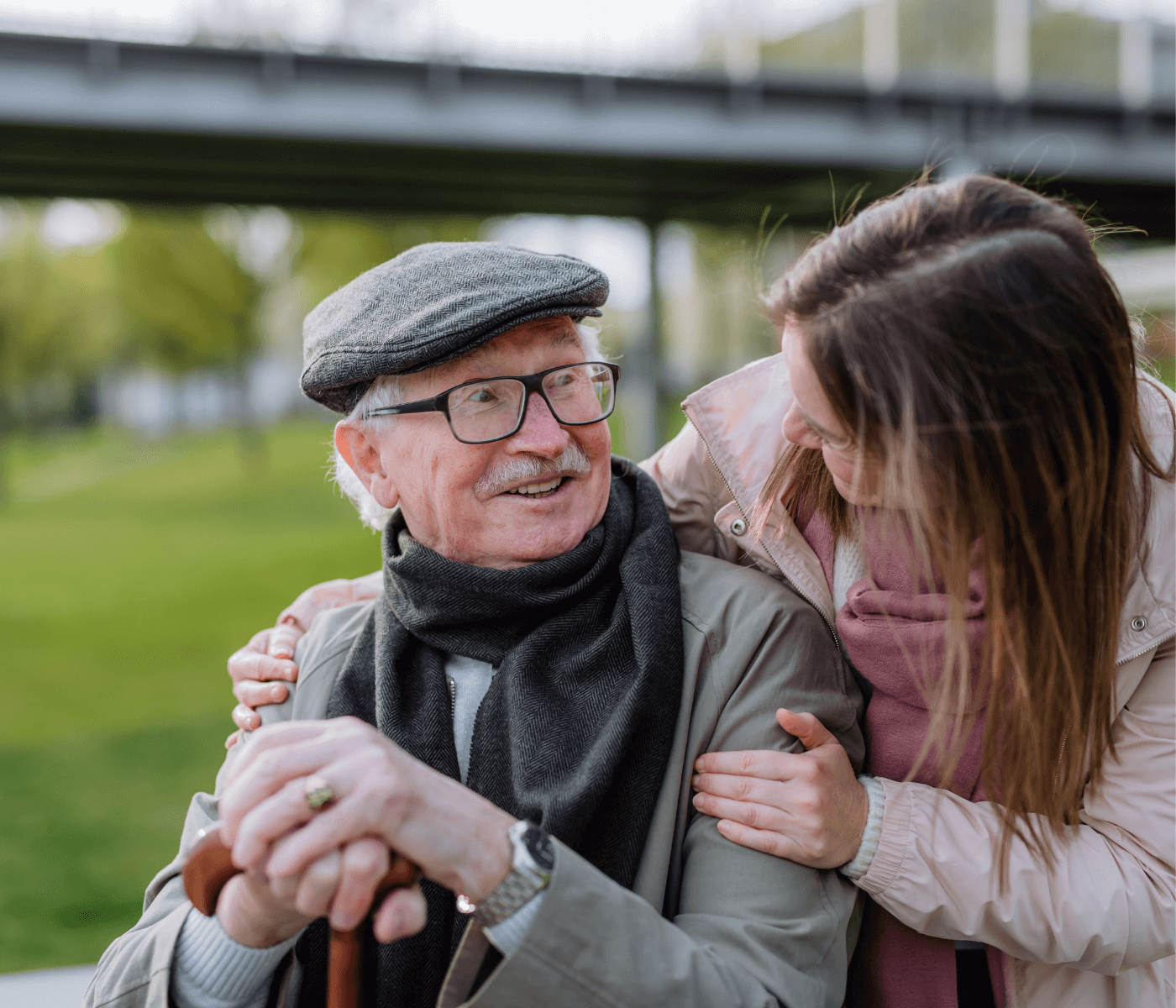 Older man with a cane smiles at a younger woman outdoors. She embraces him from behind, both wearing scarves. - Home Instead