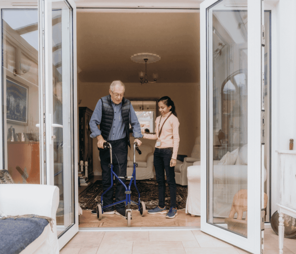Senior man using a walker is assisted by a caregiver, as they stand in an open doorway of a living room. - Home Instead