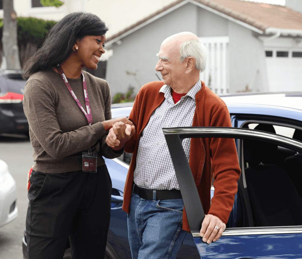 A caregiver helps an elderly man outside a car. The man smiles as he holds her hand. - Home Instead