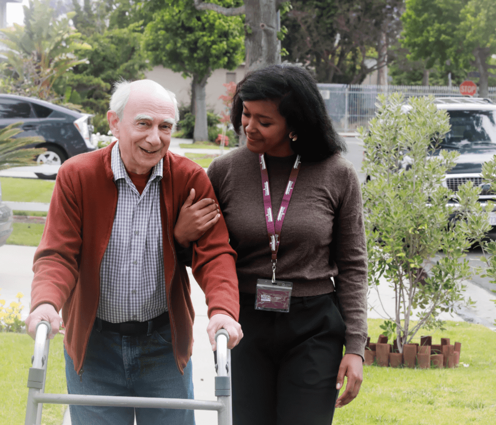 An elderly man using a walker is assisted by a caregiver as they walk outside in a garden setting. - Home Instead