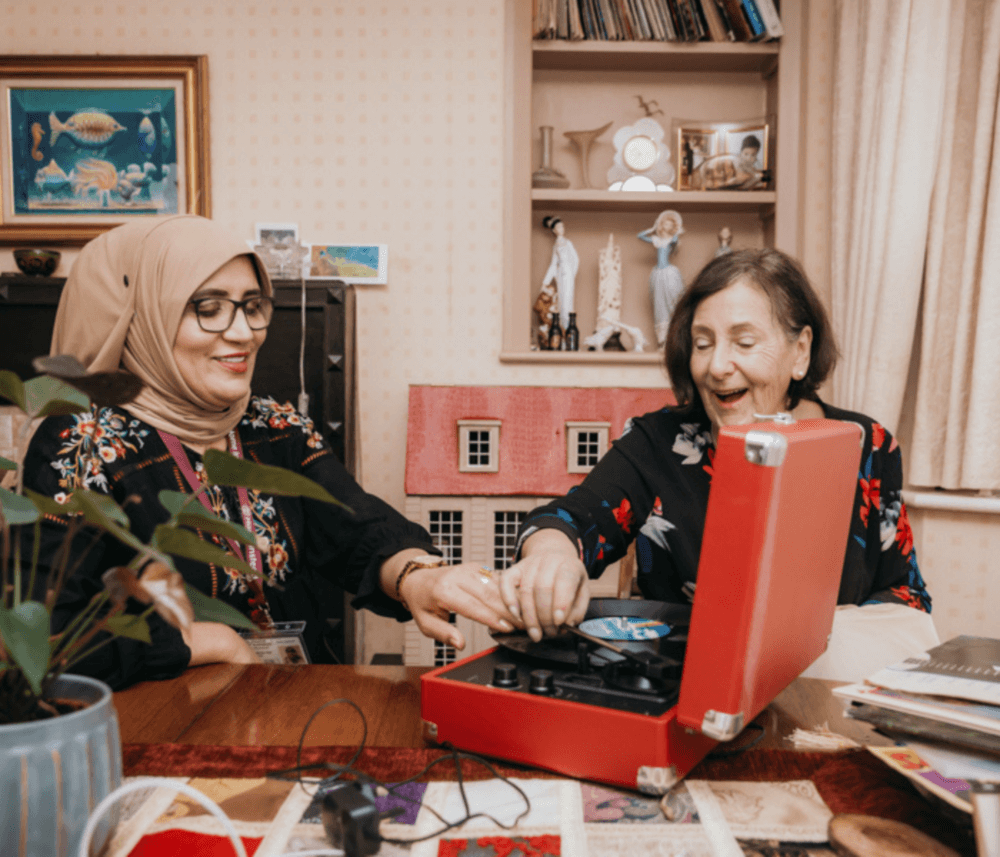 Two women smiling and interacting with a red record player on a table, in a cozy room with bookshelves and decorations. - Home Instead