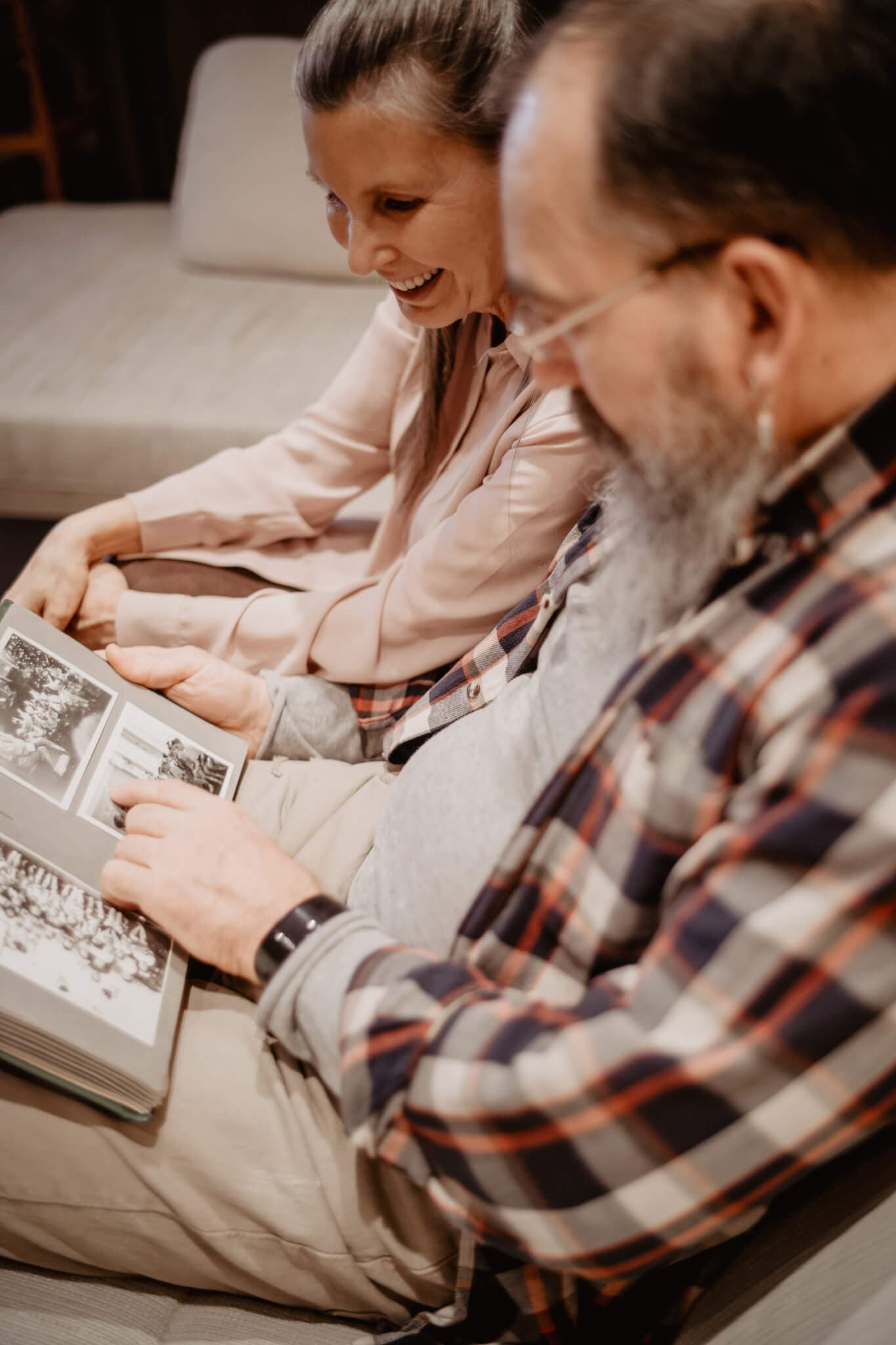 Two people sitting on a sofa, smiling and looking at an old photo album together. - Home Instead