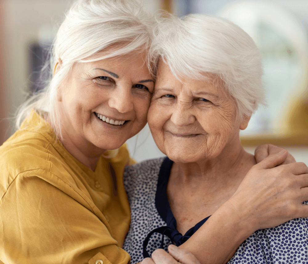 Two older women smiling and embracing, one with white hair in a yellow shirt, the other with white hair in a patterned shirt. - Home Instead