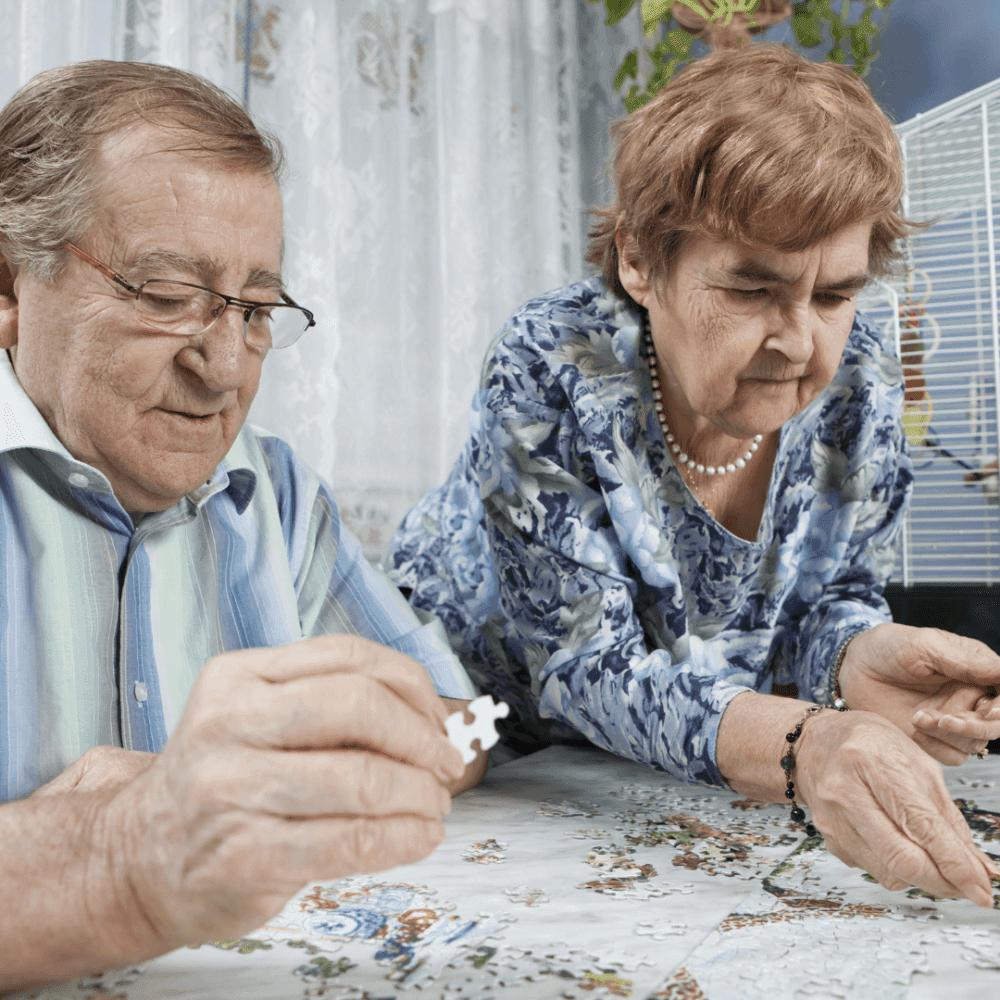 Two elderly people are focused on assembling a jigsaw puzzle at a table covered with a lace tablecloth. - Home Instead