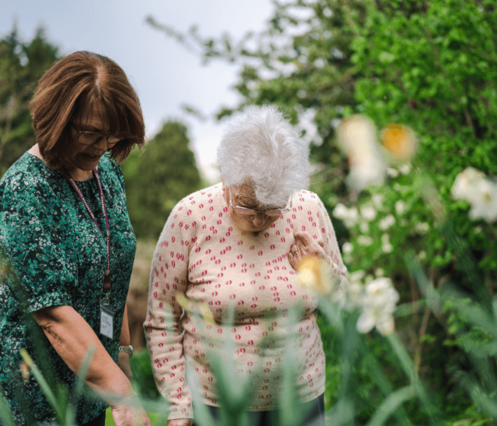 Two elderly women stand amidst lush greenery and flowers, looking down engagingly at something in the garden. - Home Instead