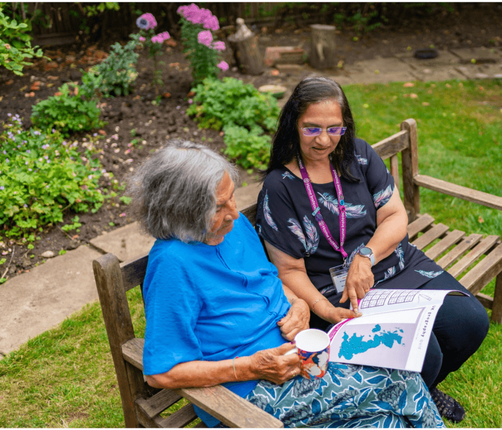 Two women sit on a bench in a garden. One shows a map to the other, who holds a cup, with flowers in the background. - Home Instead