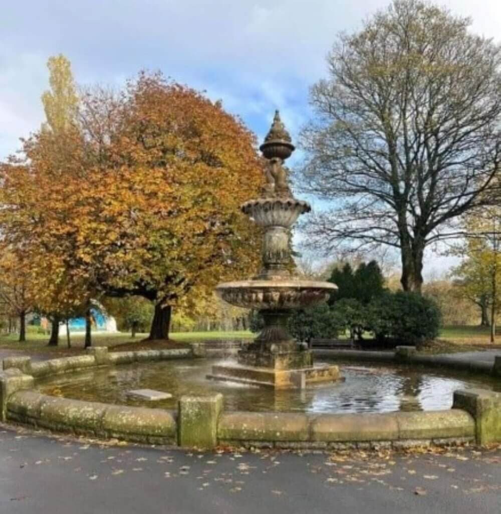 A stone fountain in a park with autumn-colored trees and a cloudy sky in the background. - Home Instead