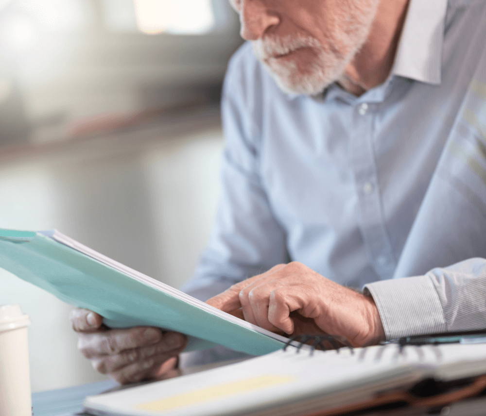Elderly man reads documents at a desk, wearing a light blue shirt. A coffee cup and papers are on the desk. - Home Instead