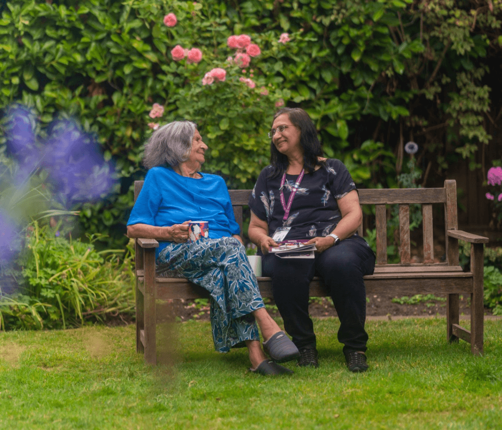 Two women sitting on a bench in a garden, smiling at each other, with flowers and greenery in the background. - Home Instead