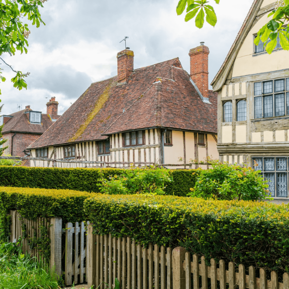 Old English houses with timber framing, a picket fence, and lush greenery under a cloudy sky. - Home Instead