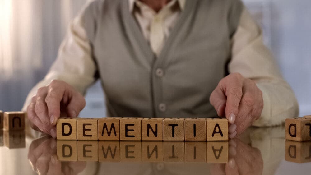 Elderly person arranging wooden blocks spelling "DEMENTIA" on a table. - Home Instead