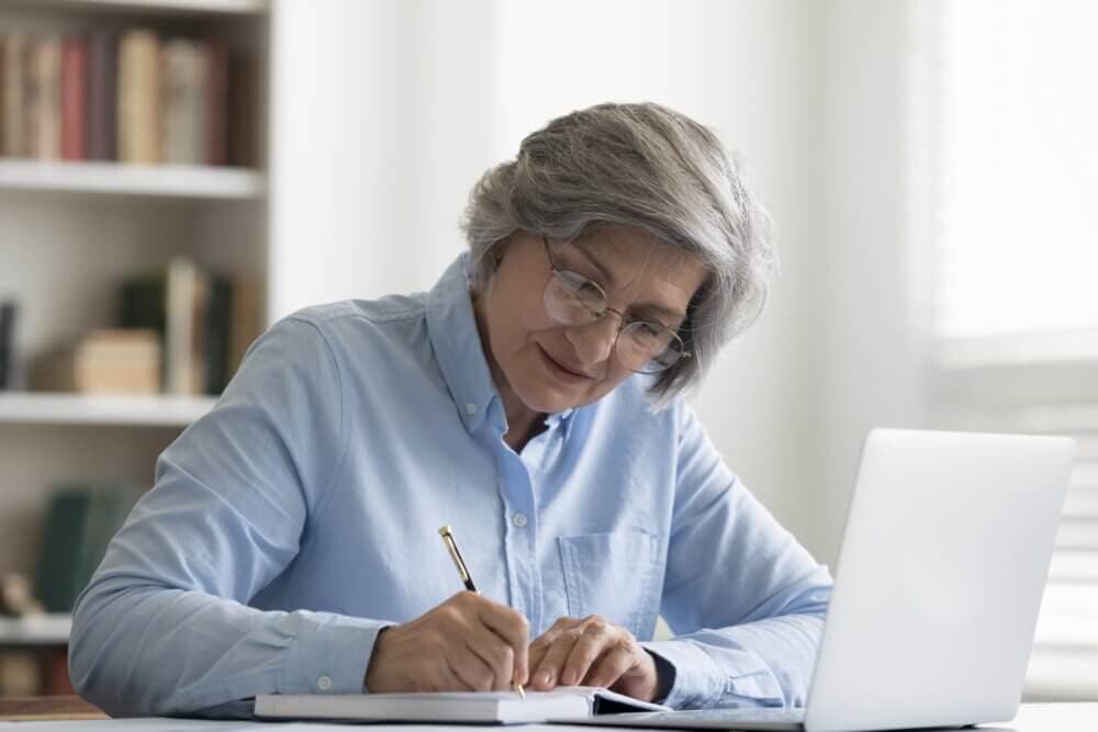 Older adult with gray hair and glasses writing in a notebook, sitting at a table with a laptop. Bookshelf in the background. - Home Instead