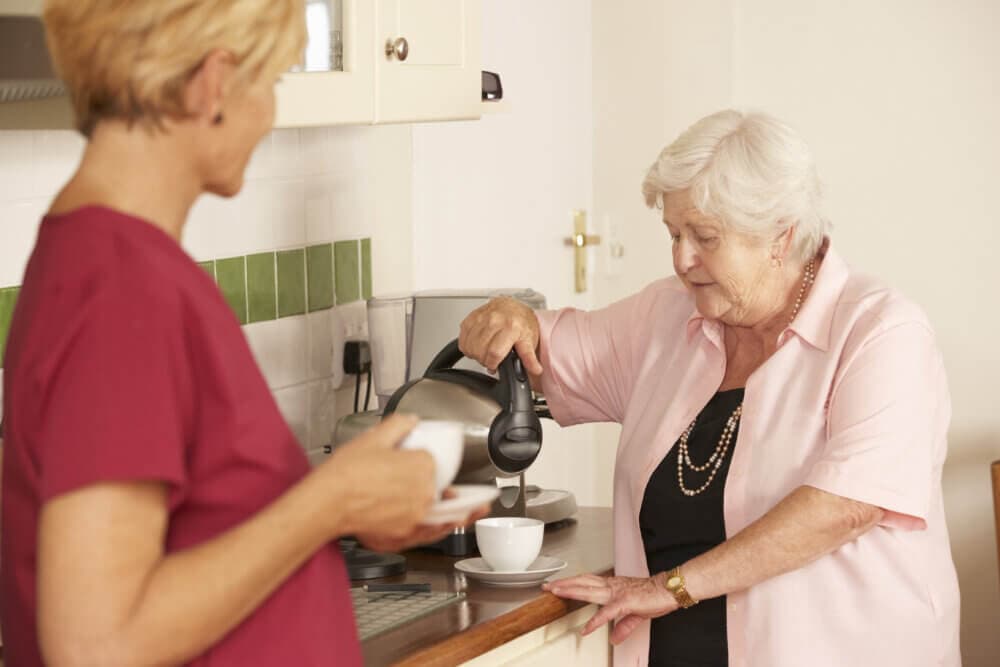 Elderly woman pours tea into a cup as a caregiver in a red uniform watches, standing beside her in the kitchen. - Home Instead