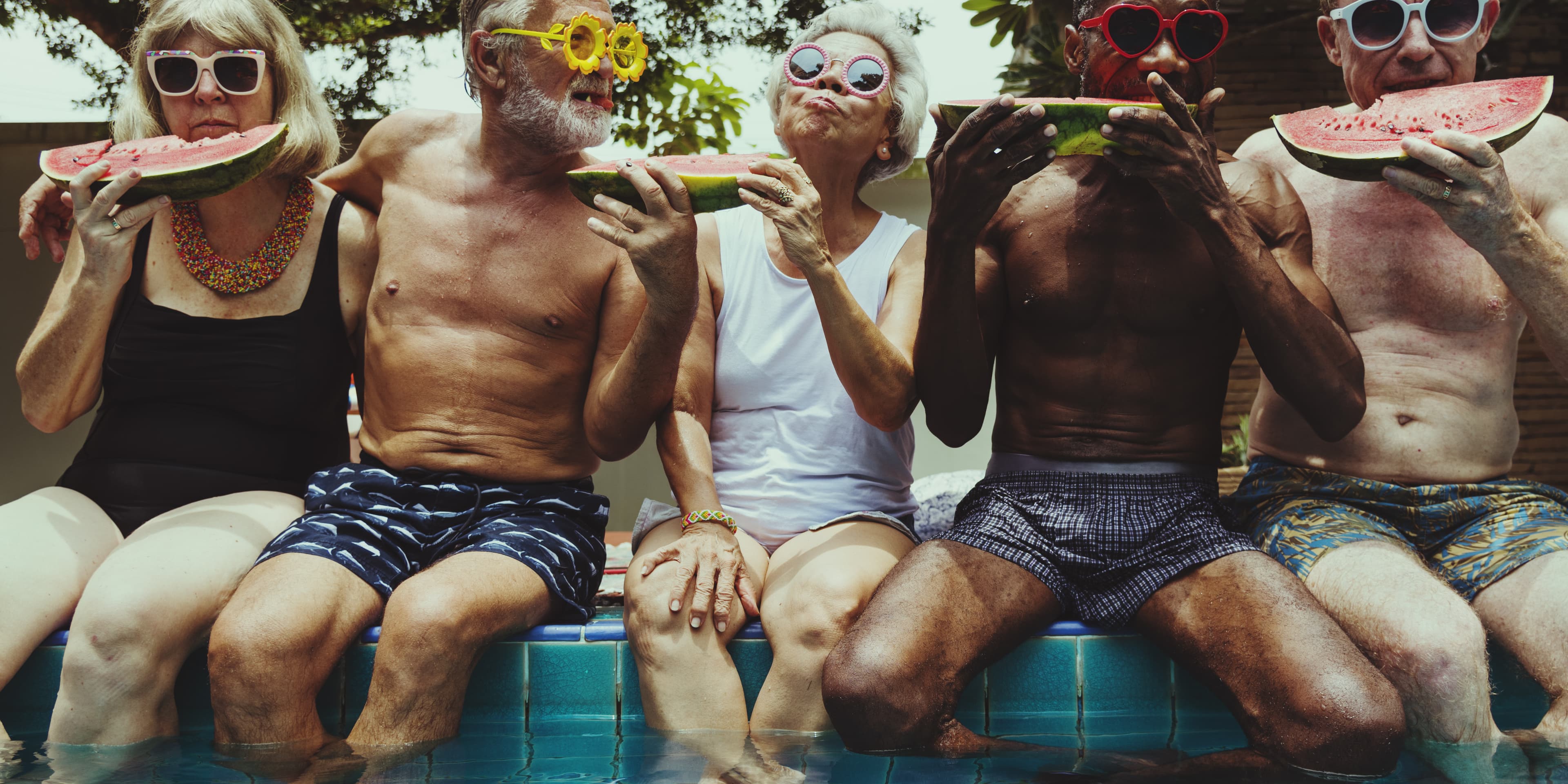 Five elderly people wearing swimsuits and colorful sunglasses sit by a pool eating watermelon slices. - Home Instead
