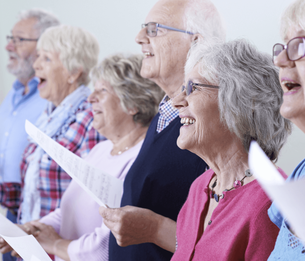 A group of elderly people singing together, holding sheets of music, and smiling. - Home Instead