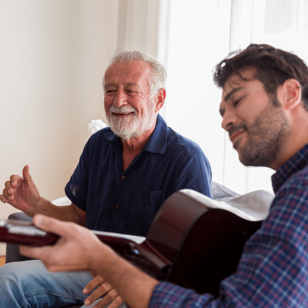 An elderly man and a younger man play the guitar together, smiling and enjoying their time. - Home Instead