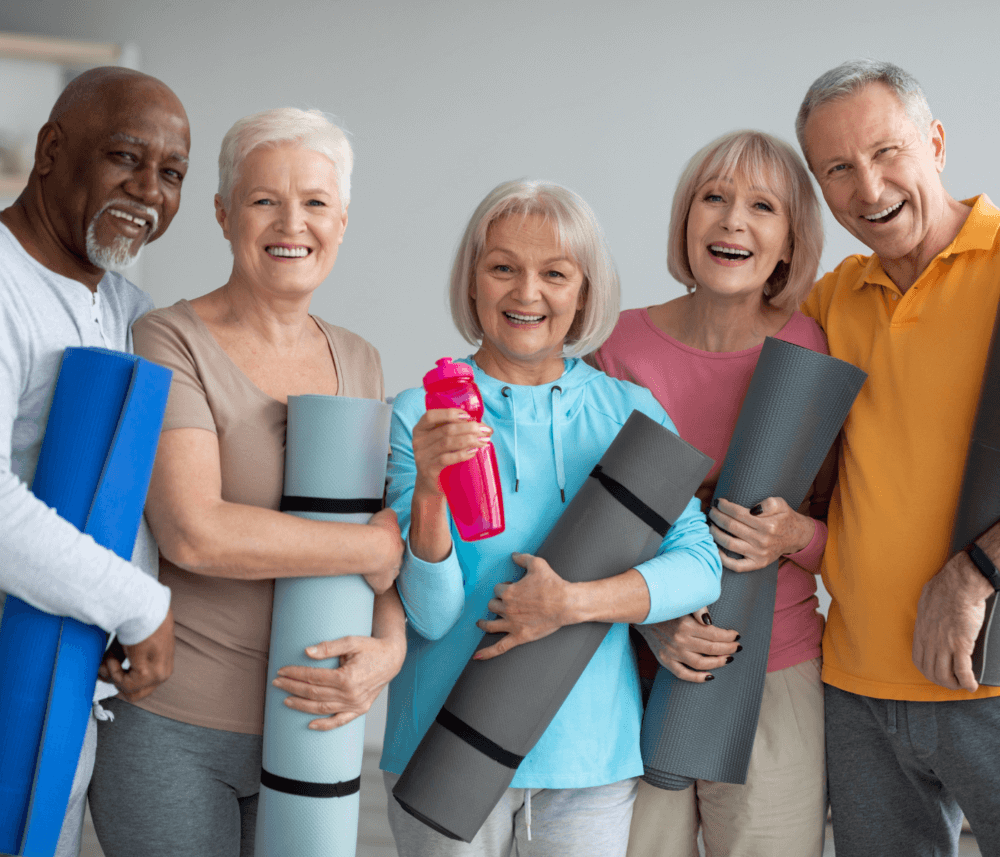 Five senior adults smiling and holding yoga mats, with one holding a pink water bottle, standing against a neutral background. - Home Instead