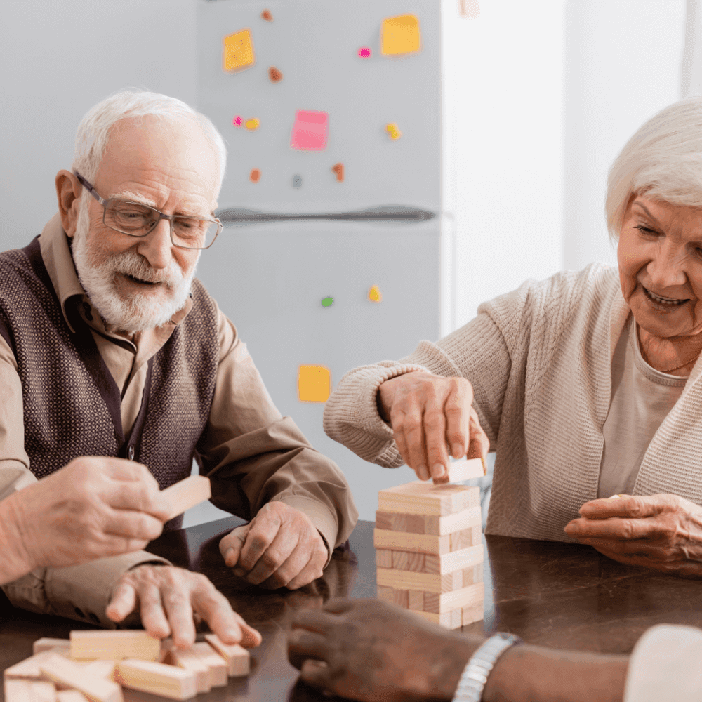 An elderly man and woman happily play a block-stacking game at a table, with a fridge covered in notes behind them. - Home Instead
