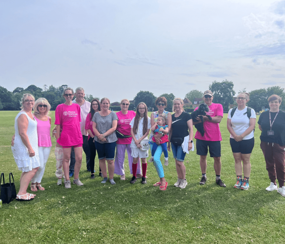 A group of people stands on a grassy field, some wearing pink shirts, posing for a group photo on a sunny day. - Home Instead