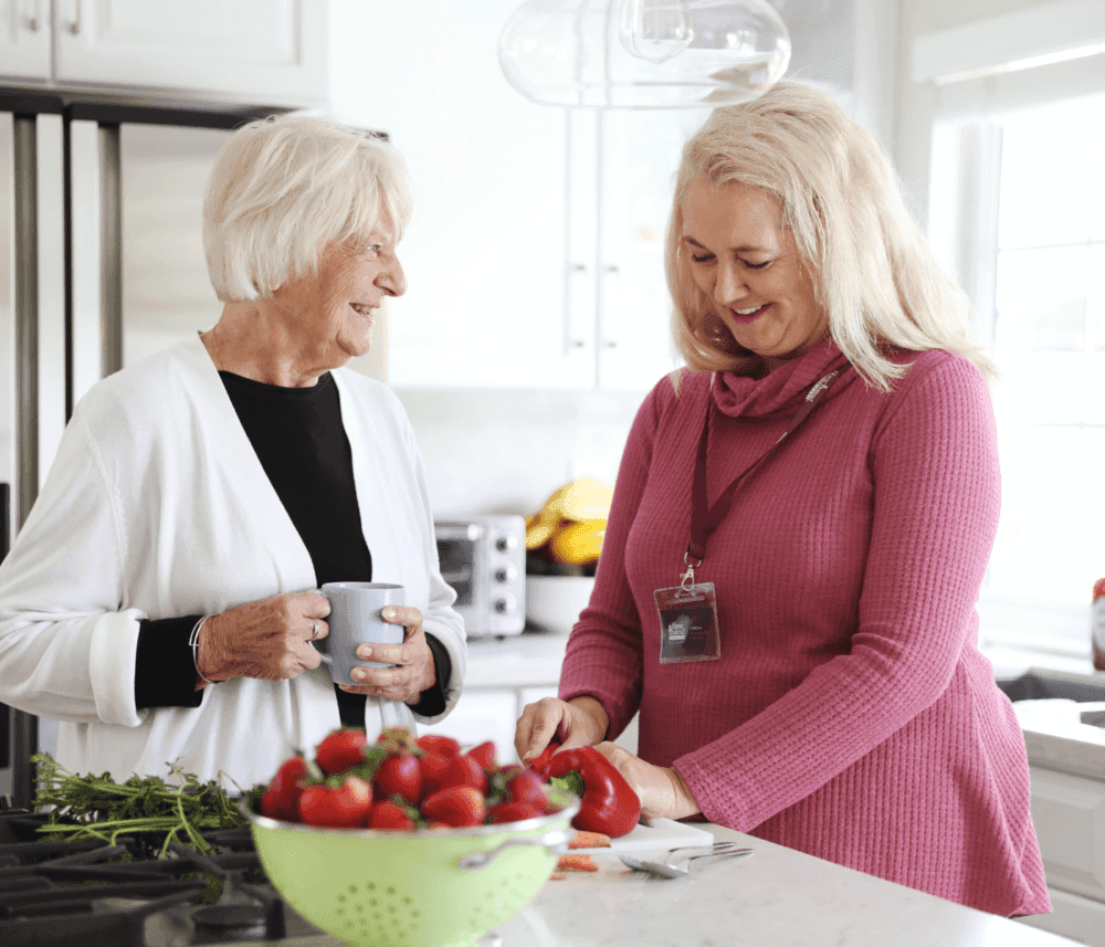 An elderly woman with a cup smiles at a woman in a pink sweater chopping vegetables in a bright kitchen. - Home Instead