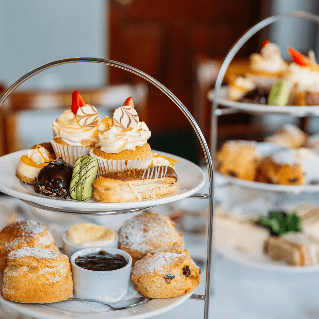 Three-tiered afternoon tea stand filled with scones, cupcakes, and assorted pastries on a white tablecloth. - Home Instead
