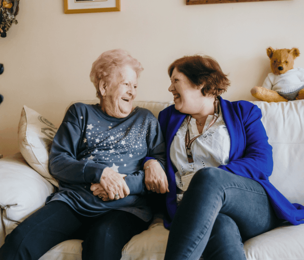 Two women smiling at each other and holding hands while sitting on a couch, with a teddy bear in the background. - Home Instead