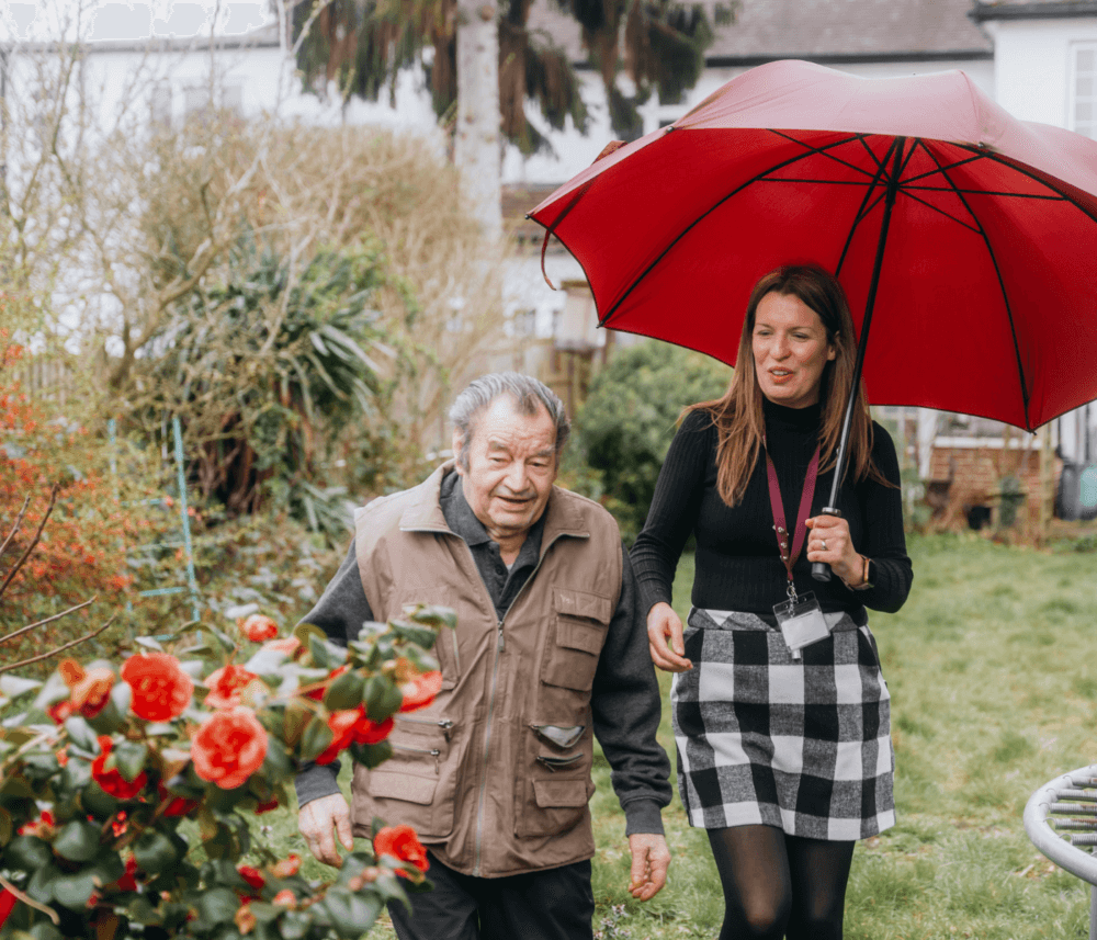 A woman holding a red umbrella walks with an elderly man in a garden with red flowers. - Home Instead