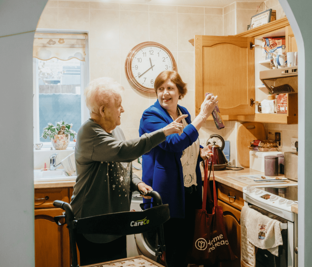 Two elderly women in a kitchen, one pointing and one reaching into a cabinet, both smiling and engaged. - Home Instead