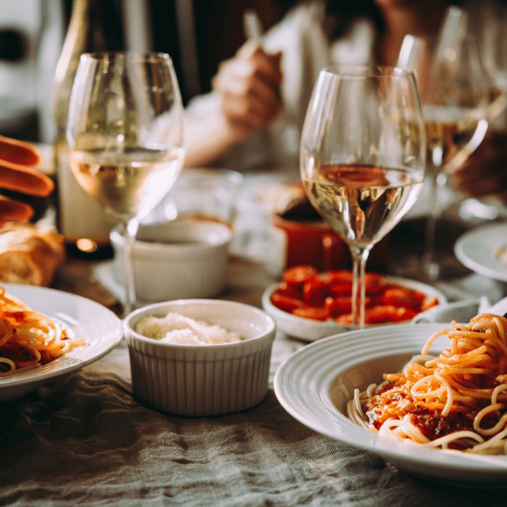Close-up of a dining table with pasta dishes, wine glasses, and various side dishes. People are partially visible in the background. - Home Instead