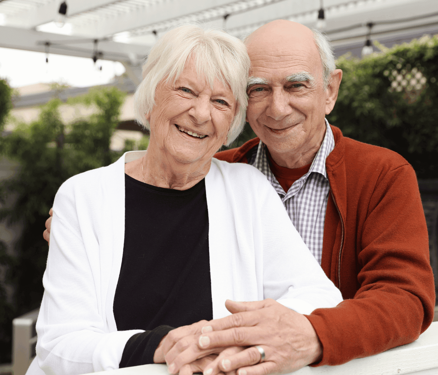 Smiling elderly couple posed together outdoors. The man is wearing a red jacket; the woman is in a white cardigan. - Home Instead