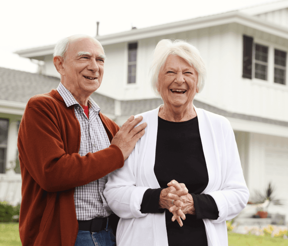 Smiling elderly man and woman standing outside a white house, both dressed in casual clothing. - Home Instead
