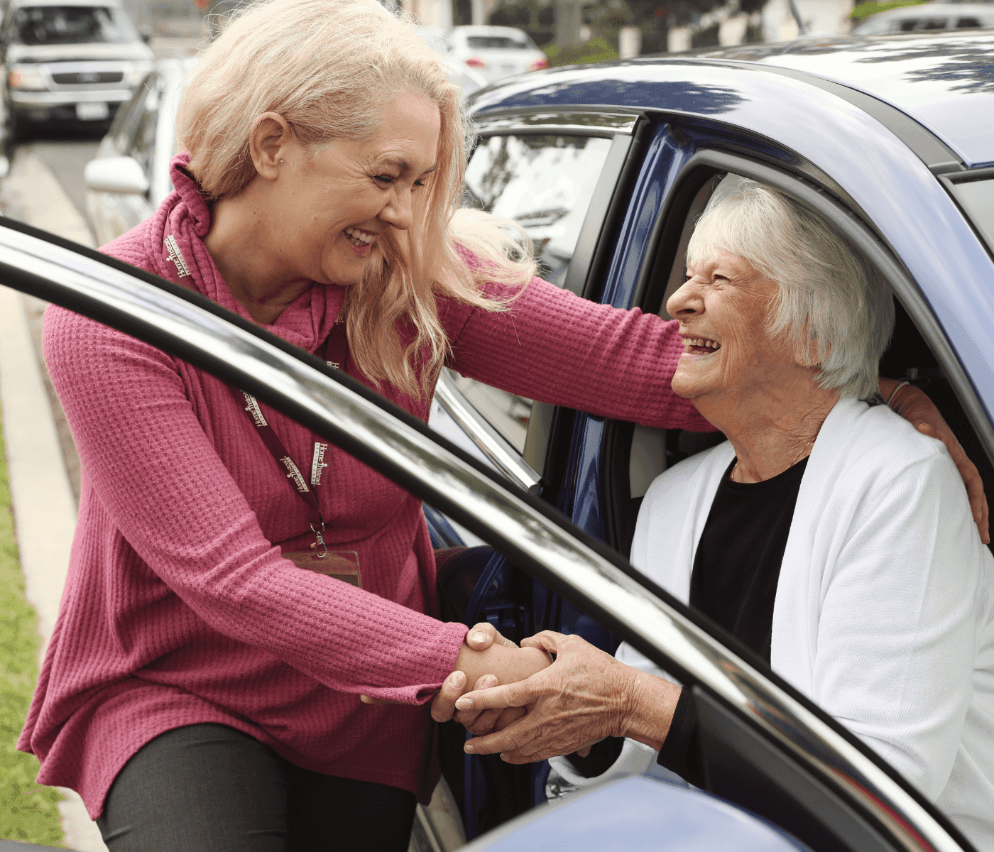 A smiling caregiver helps an elderly woman out of a car, holding her hand for support. - Home Instead