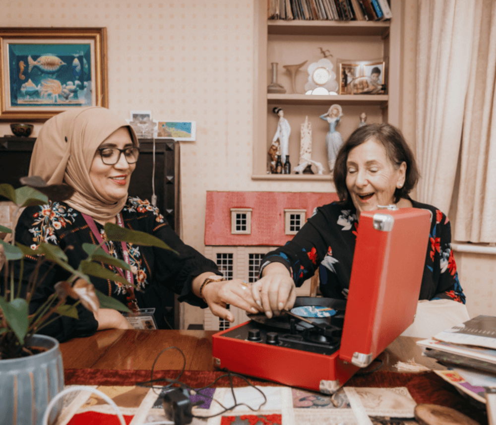 Two women, smiling and enjoying, play a vinyl record on a red turntable in a cozy room with various decorations. - Home Instead