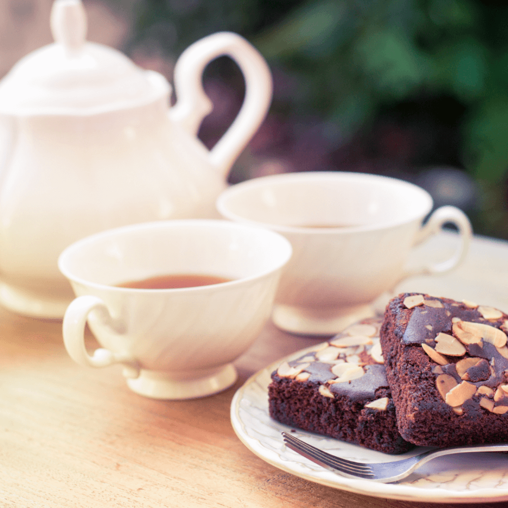 Two cups of tea and almond-topped chocolate brownies on a plate, with a white teapot in the background. - Home Instead