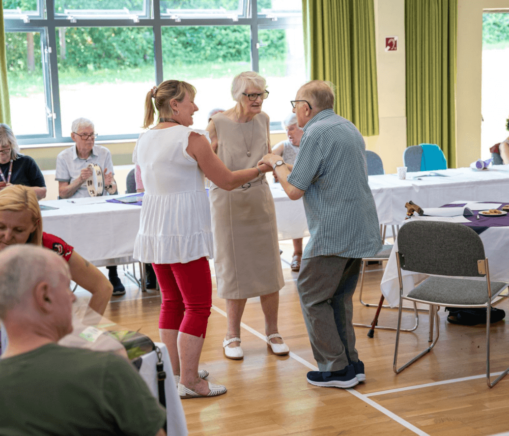 Three elderly people dancing together happily in a bright, sunlit room filled with seated attendees and tables. - Home Instead